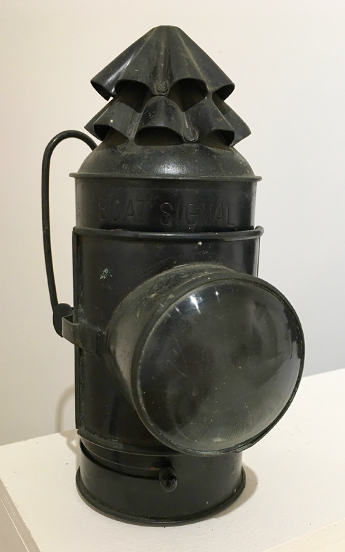 <h3>Signal Lantern</h3>
<br>
<p><i>National Marine Lamp Company</i>
<p><strong>Early twentieth century
<br>
Gift of Gabriel Latham</strong></p>
<br>
<br>
<p>This maritime lantern was used to communicate or signal between ships or boats. There is no doubt about the use of this object: large letters just above the lens spell “Boat Signal.” The lantern transmitted Morse code by using the sliding shutter located just beneath the door of the lamp.  This finger-operated shutter allowed or prevented light to shine through the thick, blown-glass bull’s eye lens. The interior contains a removable font and burner for kerosene. This hand-held lantern has double swing bail handles at the back. There is, in addition, a mounting bracket on the back. Signal lanterns were also an essential part of the equipment found in life boats.</p>