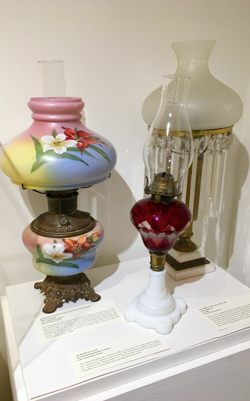 <h3>Kerosene Lamp with Hand Painted Shade</h3>
<br>
<p><i>Maker unknown</i>
<p><strong>Late nineteenth century
<br>
Gift of Charles Dean</strong></p>
<br>
<br>
<p>The thumbwheel on this kerosene lamp is marked “Made in US of America.” Other than that there are no markings, which is not unusual in glass lamps of this period. They were made in vast quantities and in innumerable designs and shapes. This example is typical of the kerosene lamps with hand-painted glass globes and shades that became ubiquitous in American houses in the second half of the nineteenth century. Today these lamps are known as chamber lamps, hurricane lamps, and “Gone with the Wind” lamps.</p>
<br>
<br>
<hr>
<br>
<br>
<h3>Kerosene Lamp</h3>
<br>
<p><i>Scovill Manufacturing Company</i>
<p><strong>Late nineteenth century
<br>
Gift of George R. Latham</strong></p>
<br>
<br>
<p>The white milk glass base contrasts vividly with the red of the etched cranberry glass top, and they are joined by a brass fitting. The thumbwheel is marked “Scovill Mfg Co” and the burner “Queen Anne No 2.” Scovill made the burners for many different manufacturers, so probably did not produce the lamp itself.</p>
<br>
<br>
<hr>
<br>
<br>
<h3>Sinumbra Type Argand Lamp</h3>
<br>
<p><i>Maker unknown</i>
<p><strong>Ca. 1840
<br>
Gift of Alice Pratt Fey</strong></p>
<br>
<br>
<p>This is a type of argand lamp called a sinumbra lamp. An argand lamp (developed by a Swiss inventor named Aime Argand) had a tubular wick which gave a bright and smokeless flame and is considered revolutionary in the development of lighting. A sinumbra lamp improved on the basic argand lamp by eliminating the cast shadow. The slope of the reservoir for the fuel and the shape of the glass shade both are factors which help eliminate the shadow. This example in bronze and marble with a glass shade and dangling prisms was electrified in the twentieth century.</p>